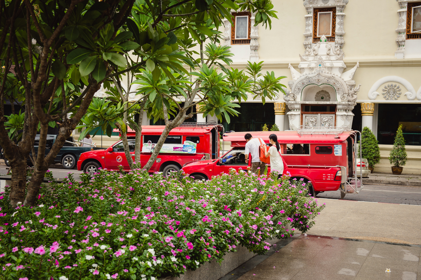 Passengers getting into a red Songthaew truck parked near colorful flowers.